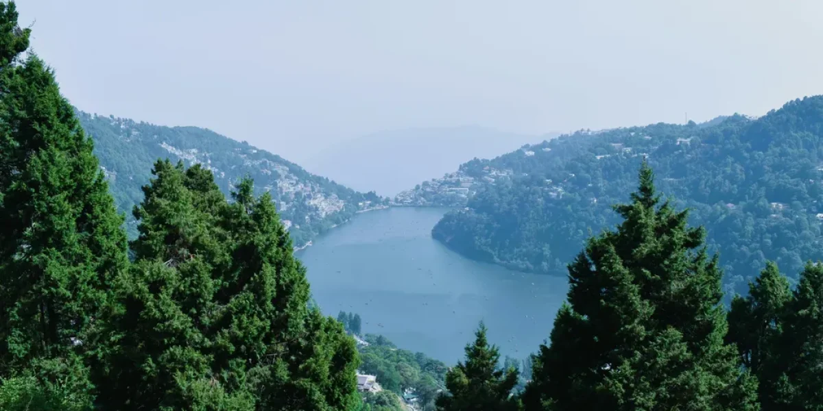 A serene view of Nainital Lake surrounded by lush green hills and pine trees, with the misty mountains fading in the distance — a perfect glimpse of the natural beauty of Nainital.