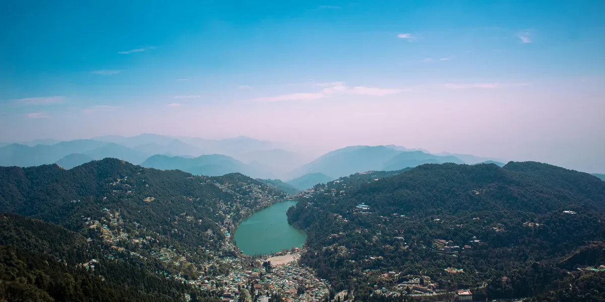 Aerial view of Nainital town with Naini Lake surrounded by lush green hills and distant Himalayan ranges under a clear blue sky.